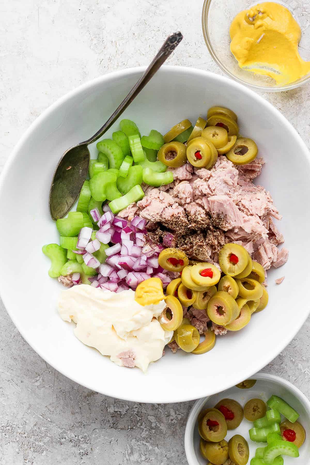 Healthy tuna salad with olives ingredients in a bowl before mixing with a spoon next to a small bowl of mustard, olives, and celery.