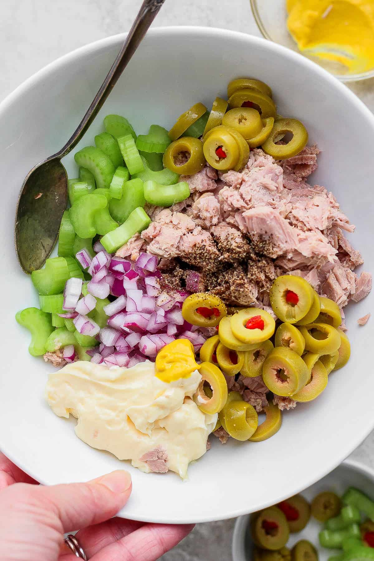 Healthy tuna salad with olives ingredients in a white bowl before being mixed with a spoon sticking out the side.