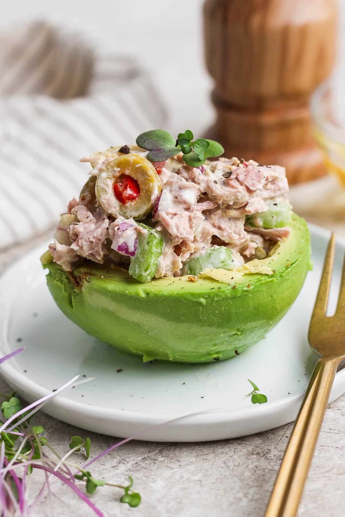 A healthy tuna salad with olives in half of an avocado on a plate with a gold fork.