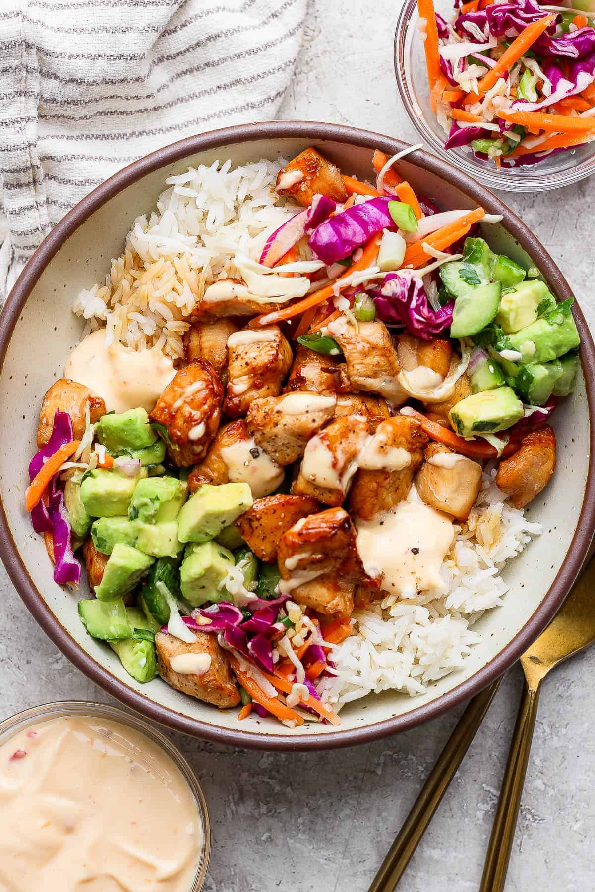 A fully assembled bang bang chicken bowl next to a small dish of bang bang sauce, another bowl of slaw, and gold silverware.
