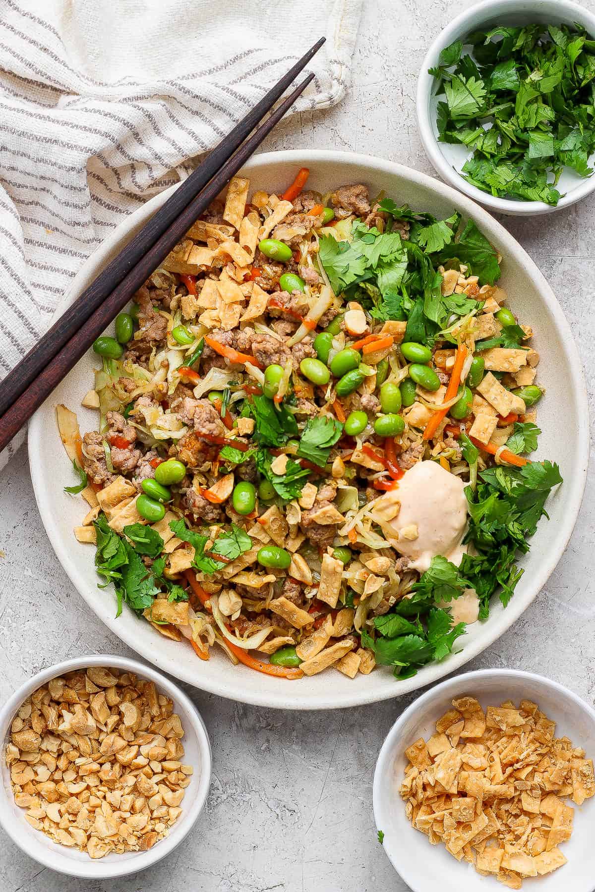 A fully assembled Blackstone egg roll in a bowl with chopsticks on the side and small bowls of cilantro, peanuts, and wonton strips next to it.