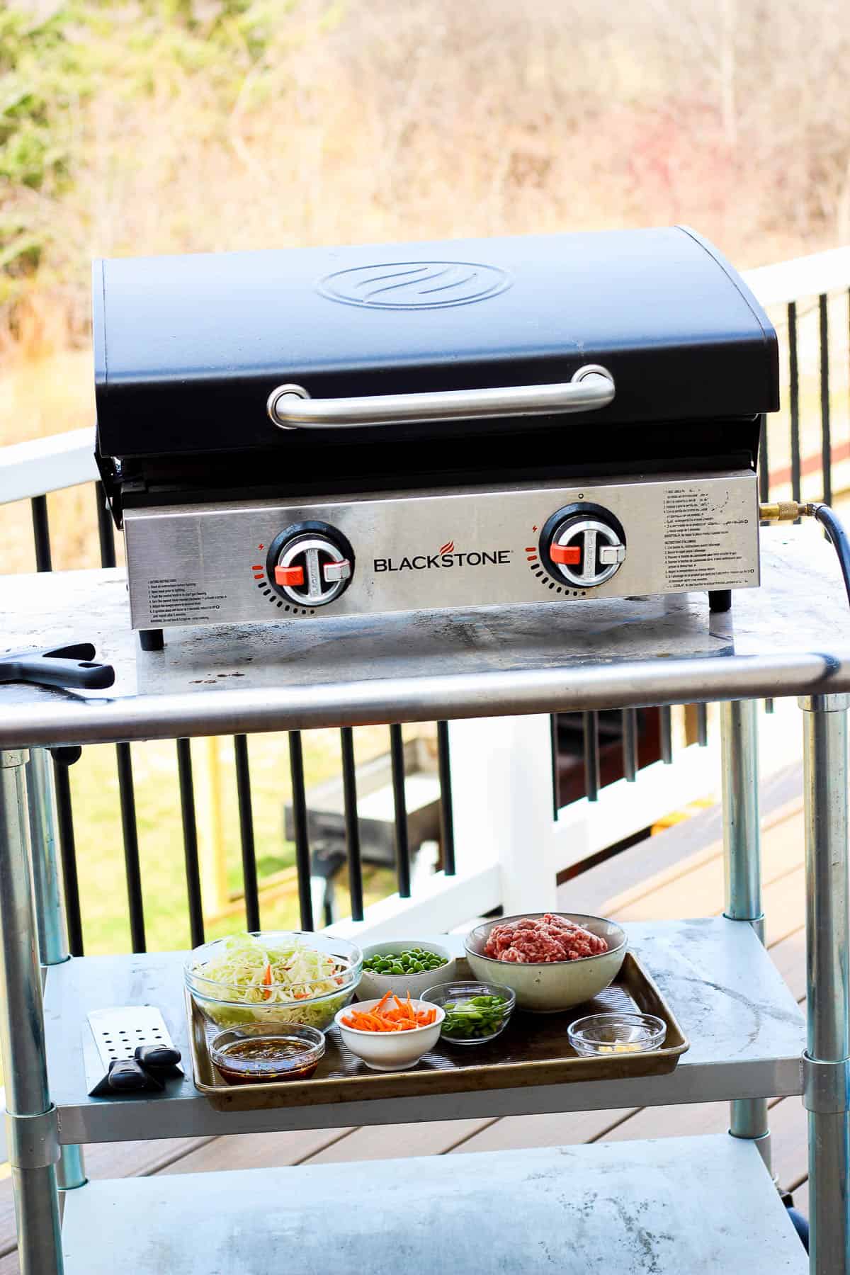 A Blackstone griddle on a metal cart with the ingredients for blackstone egg roll in a bowl on a sheet pan below it.