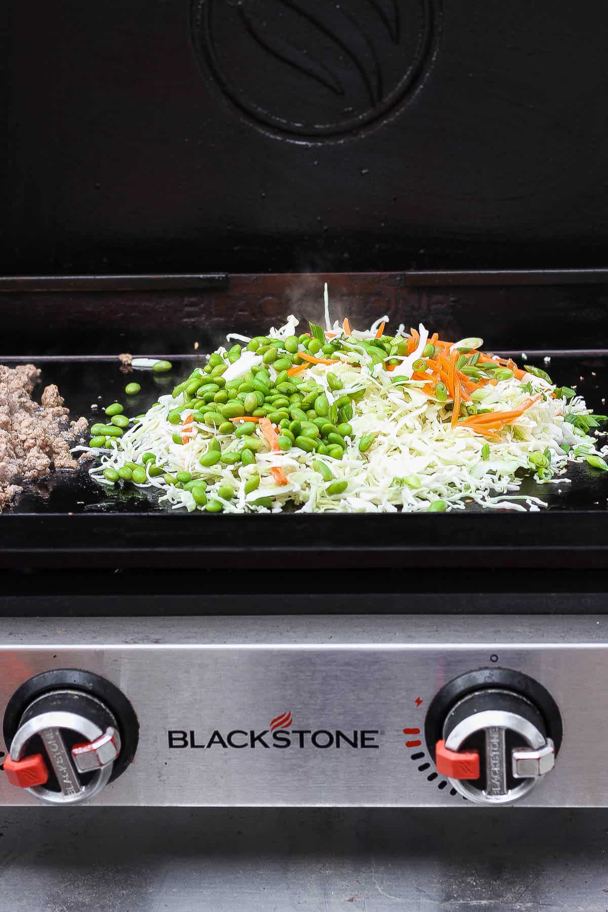 Coleslaw mix, edamame, carrots, and green onions in the middle of a Blackstone griddle with cooked pork off to the side to make Blackstone egg roll in a bowl.