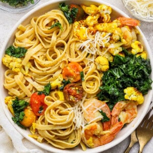 Turmeric pasta with shrimp and cauliflower in a large white bowl next to gold silverware.