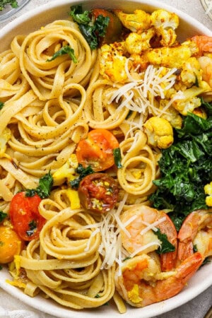 Turmeric pasta with shrimp and cauliflower in a large white bowl next to gold silverware.