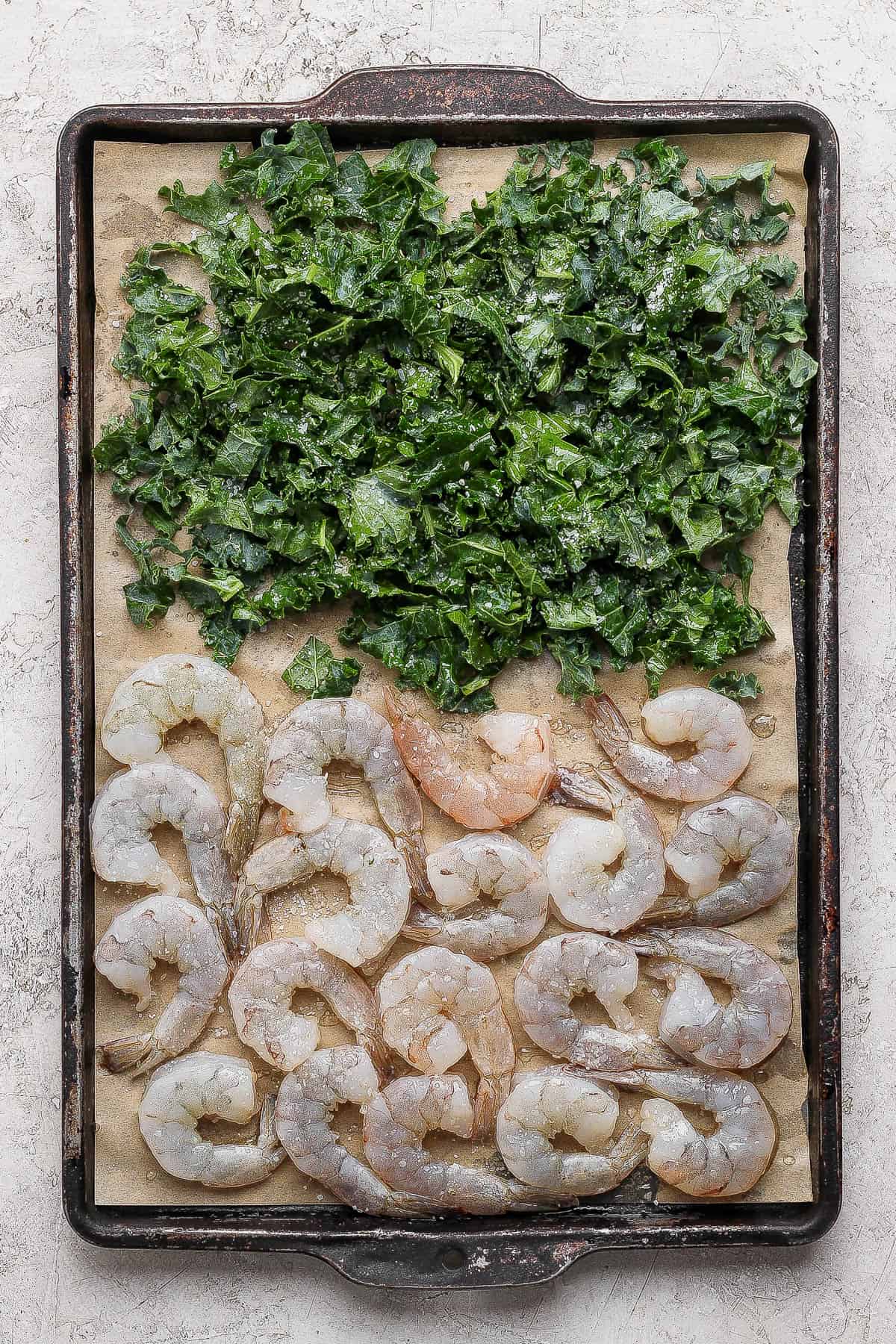 Shrimp and kale on a parchment-lined sheet pan after being tossed in olive oil and salt and before going in the oven to cook.