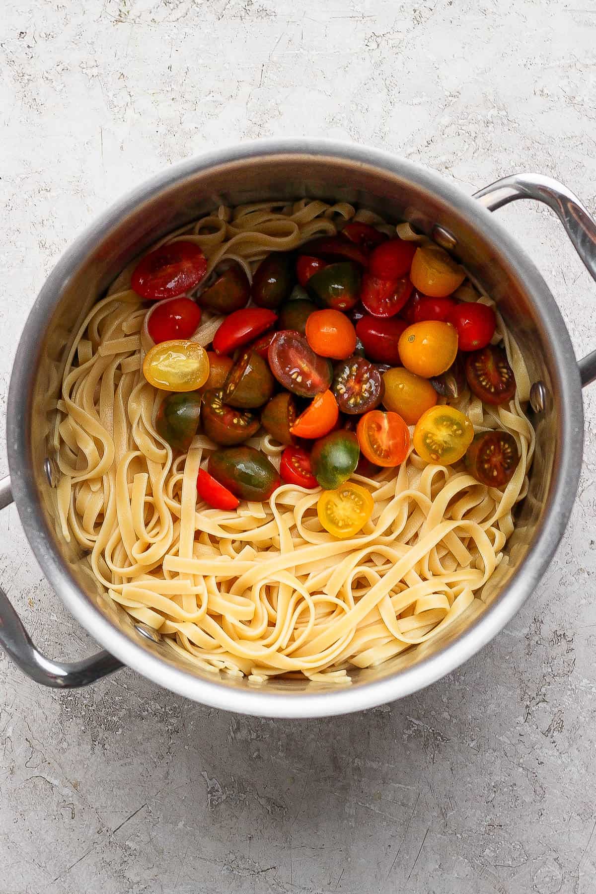 Cooked fettuccine noodles in a large metal pot with halved cherry tomatoes.