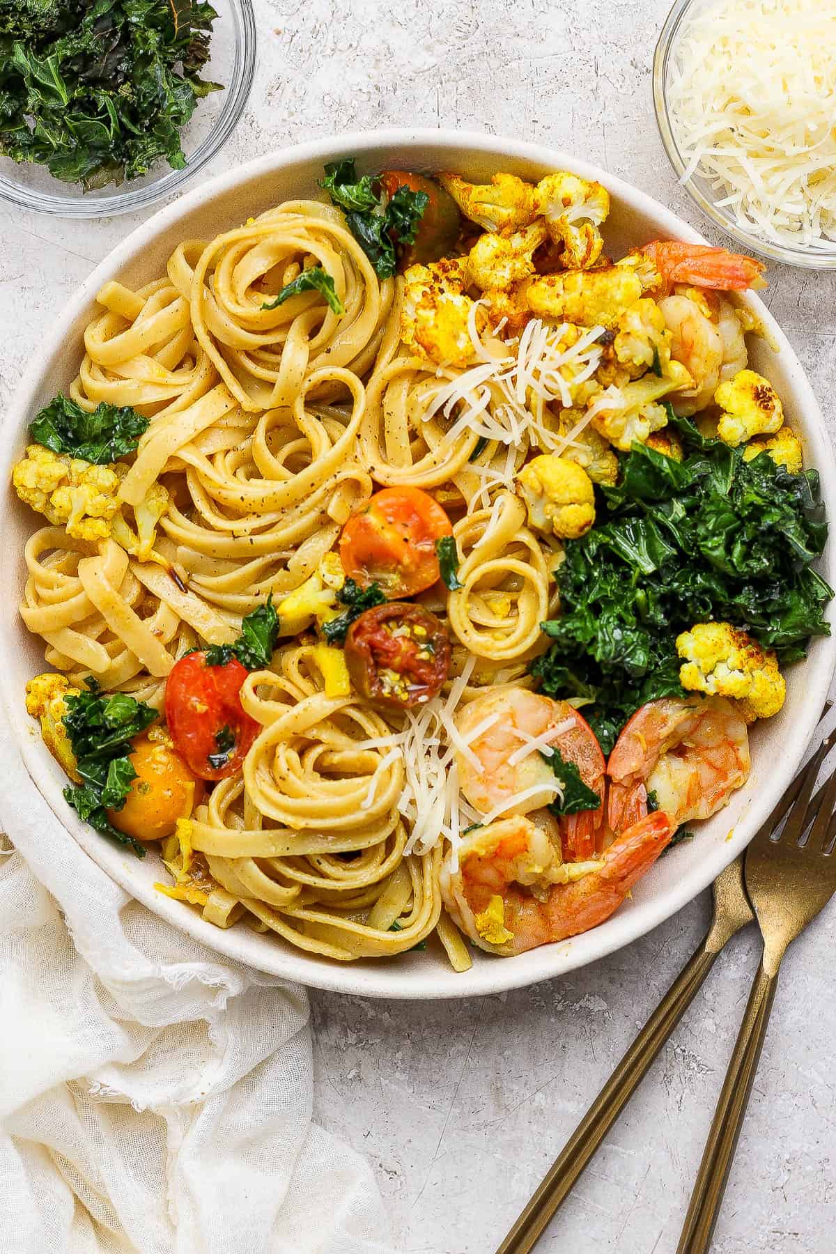 Turmeric pasta with shrimp and cauliflower in a bowl with parmesan cheese on top and next to gold silverware and small bowls of parmesan and kale.