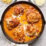 Creamy tomato chicken thighs fully cooked in a skillet next to small bowls of parmesan cheese and noodles.