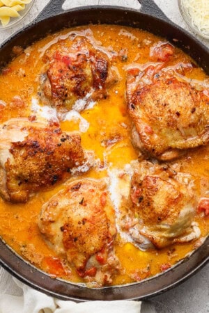 Creamy tomato chicken thighs fully cooked in a skillet next to small bowls of parmesan cheese and noodles.