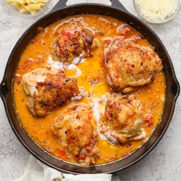 Creamy tomato chicken thighs fully cooked in a skillet next to small bowls of parmesan cheese and noodles.