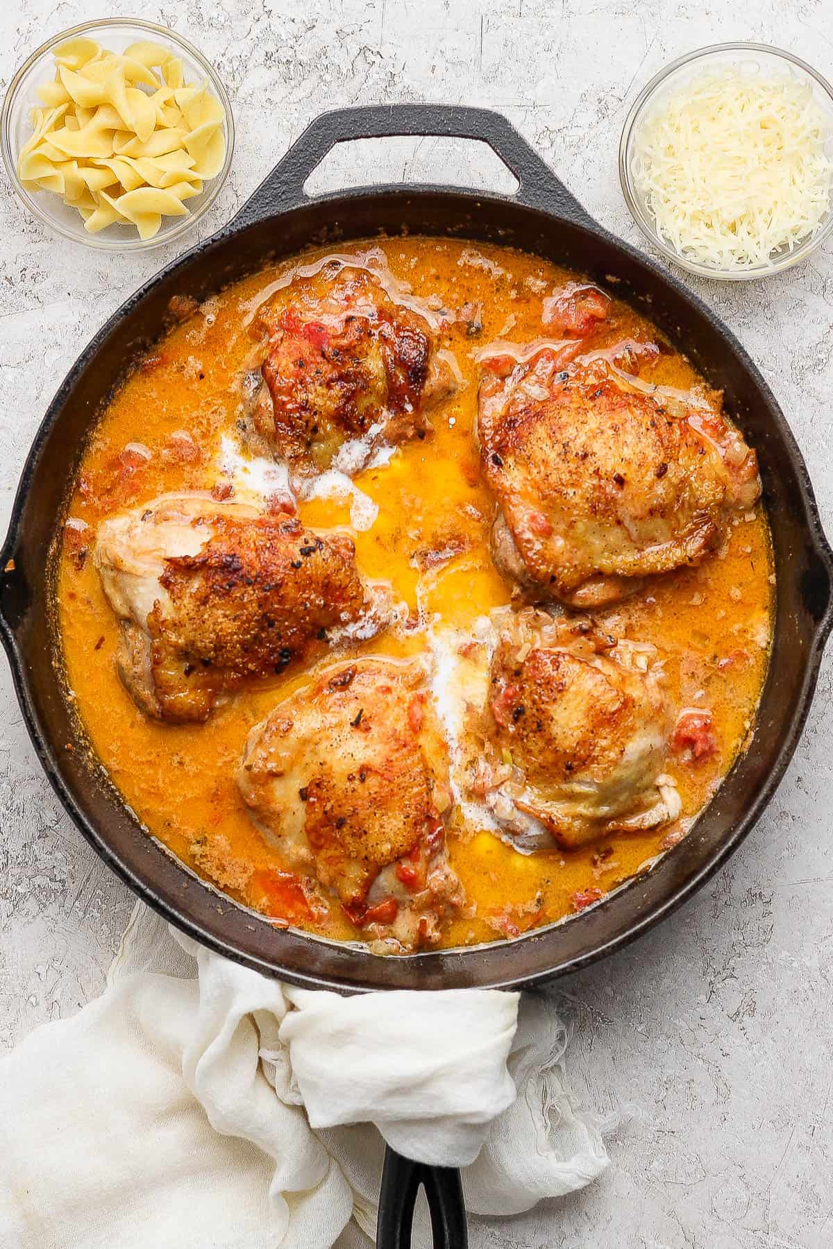 Fully cooked creamy tomato chicken thighs in a skillet next to small bowls of egg noodles and parmesan cheese.