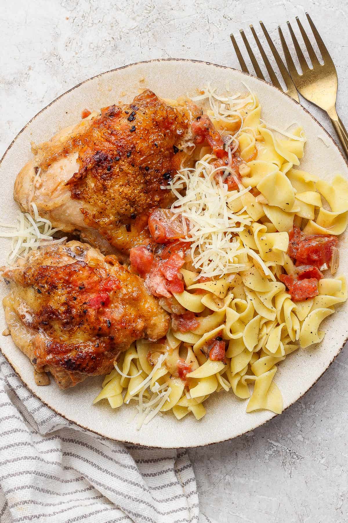 Creamy tomato chicken thighs served on a plate over egg noodles with parmesan cheese on top.