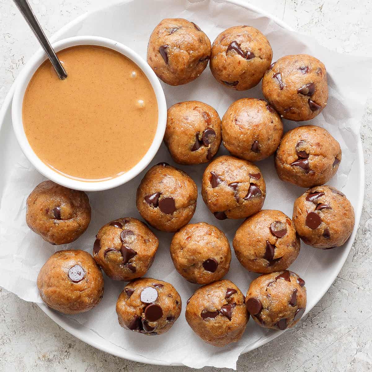 A plate filled with homemade chocolate chip cooke dough bites with a bowl of peanut butter with a spoon sticking out.