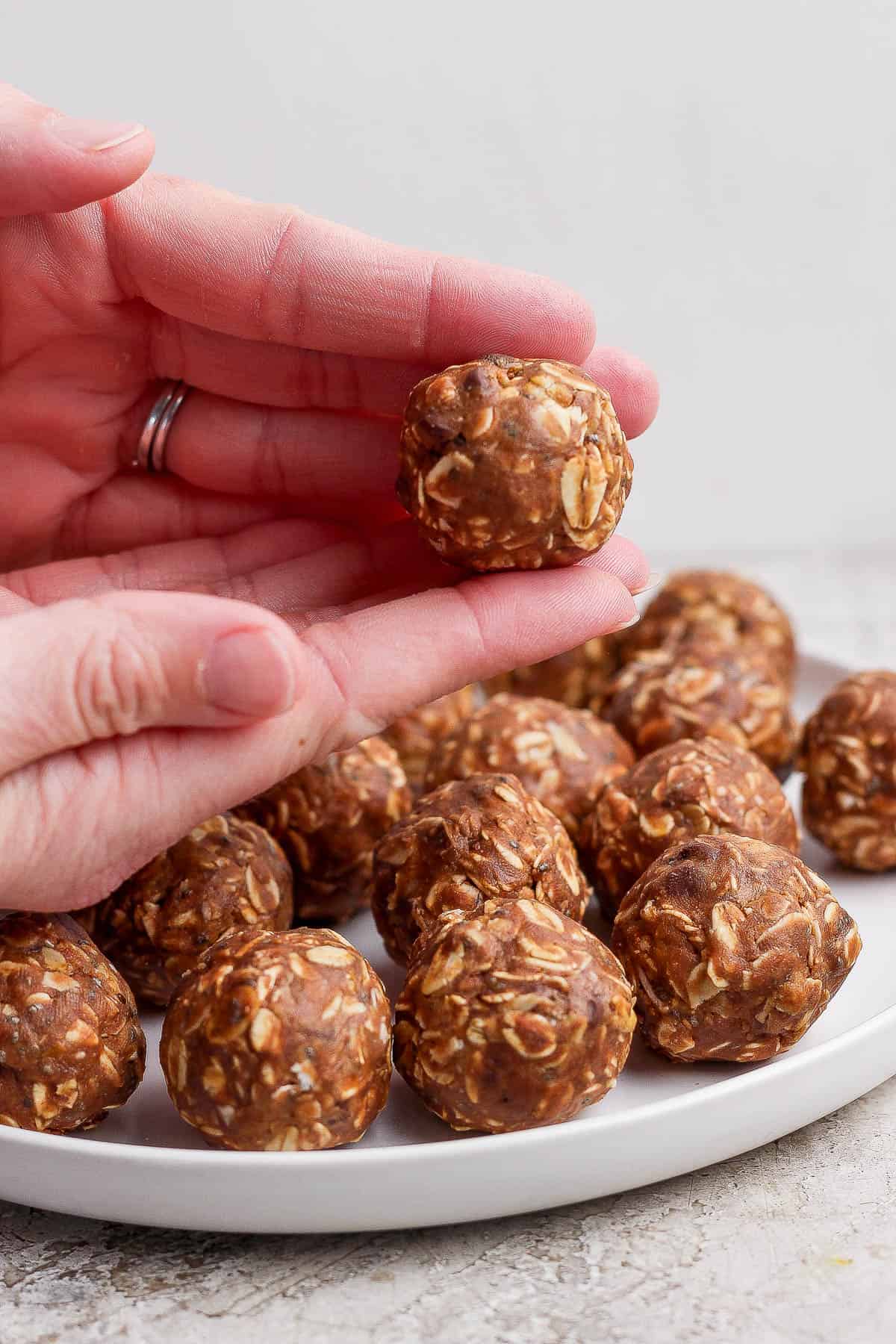Several chocolate peanut butter energy balls on a white plate with two hand rolling one more above them.