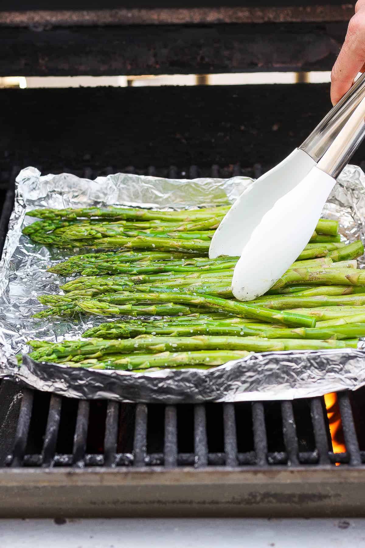 Grilled asparagus in foil on the grill and being rotated with a pair of tongs.