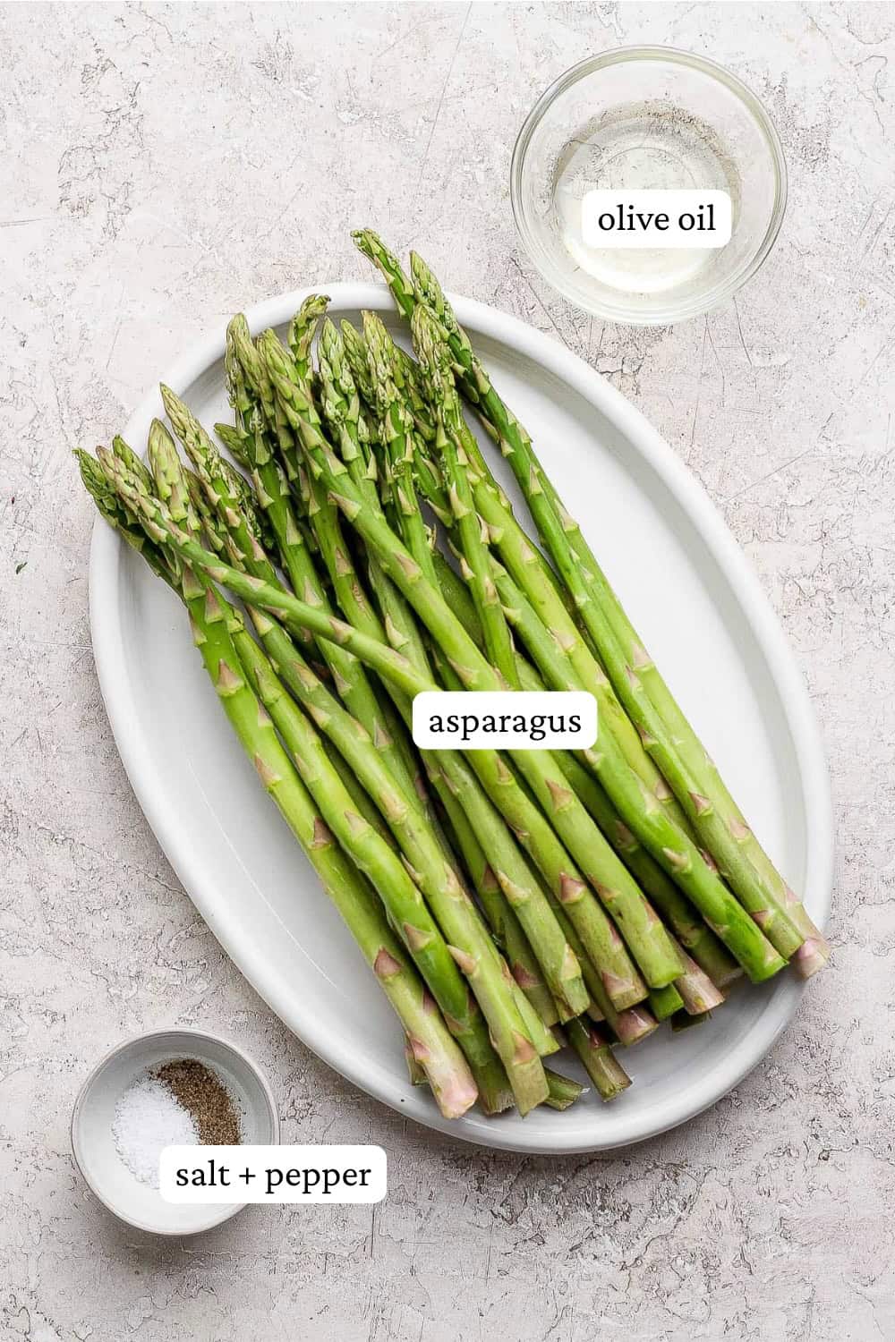 Asparagus on a white plate next to bowls of oil and salt and pepper with labels.