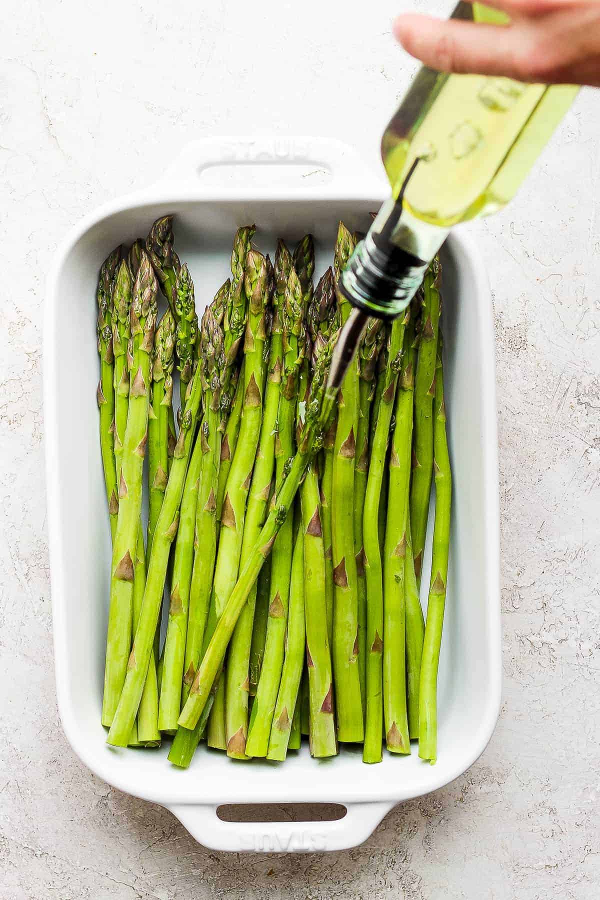 Trimmed asparagus in a white baking dish with olive oil being poured on them before grilling.
