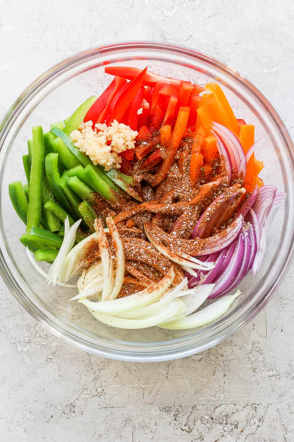 Fresh peppers, onions, garlic, and fajita seasoning in a glass bowl before being mixed and cooked.