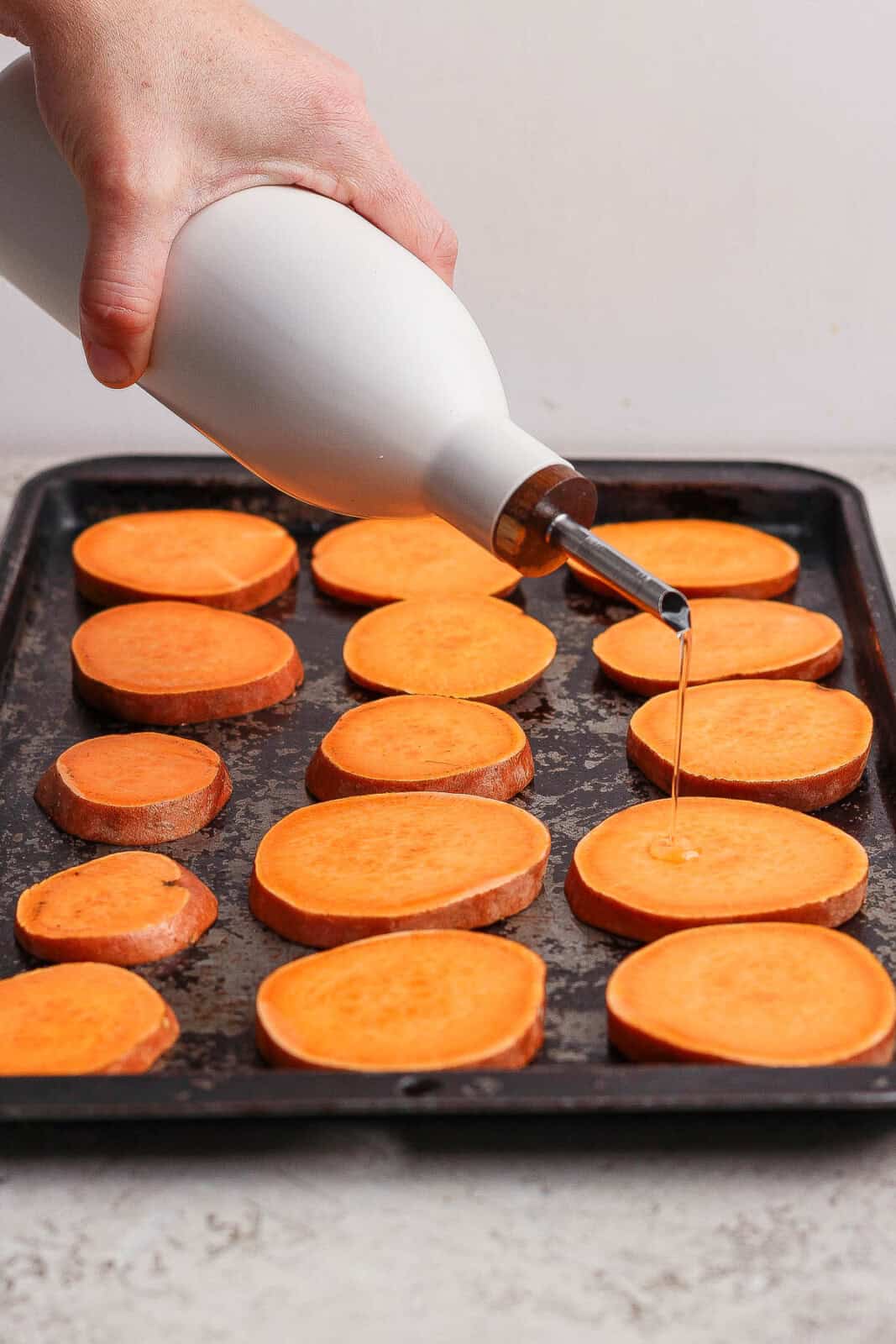 Cut sweet potatoes on baking sheet with someone pouring oil on them.