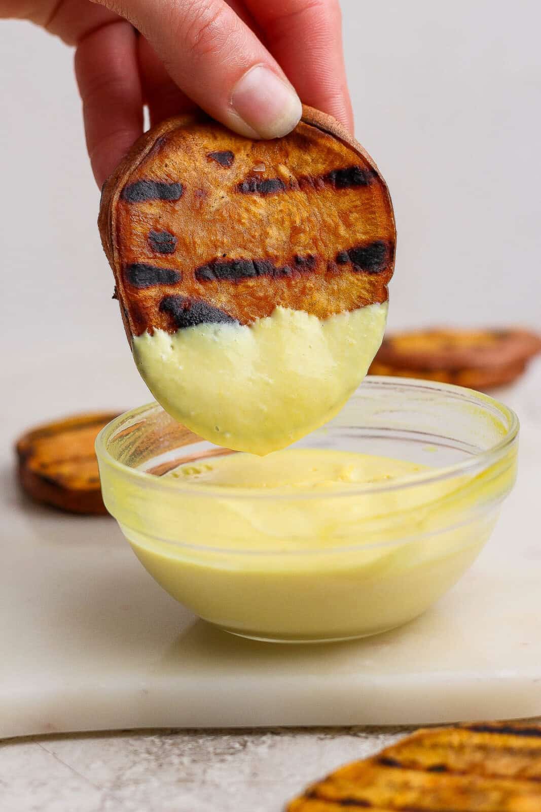 Someone holding a slice of grilled sweet potato and dipping it into some mustard mayo.