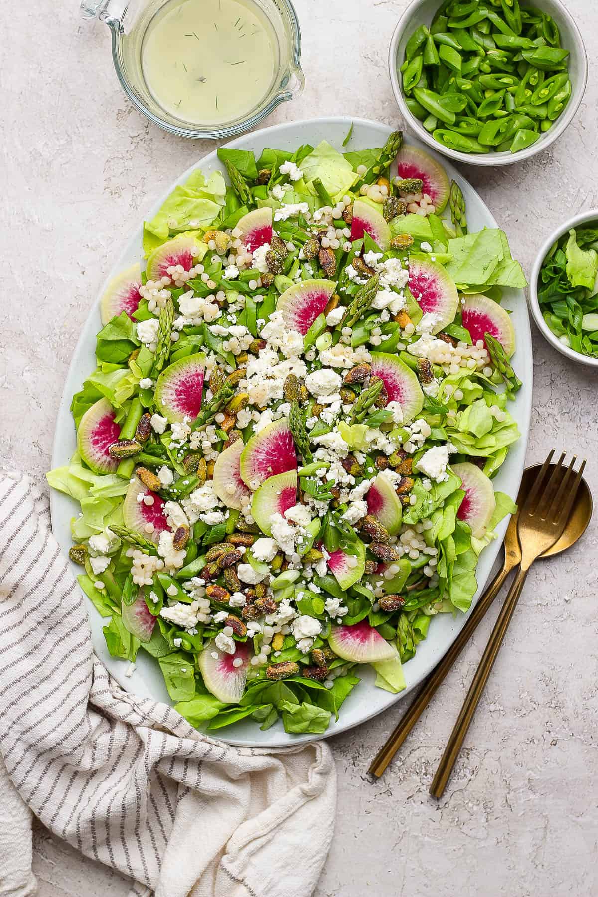 A fully assembled spring salad with couscous and peas next to small dishes of lettuce, chopped peas, and vinaigrette.