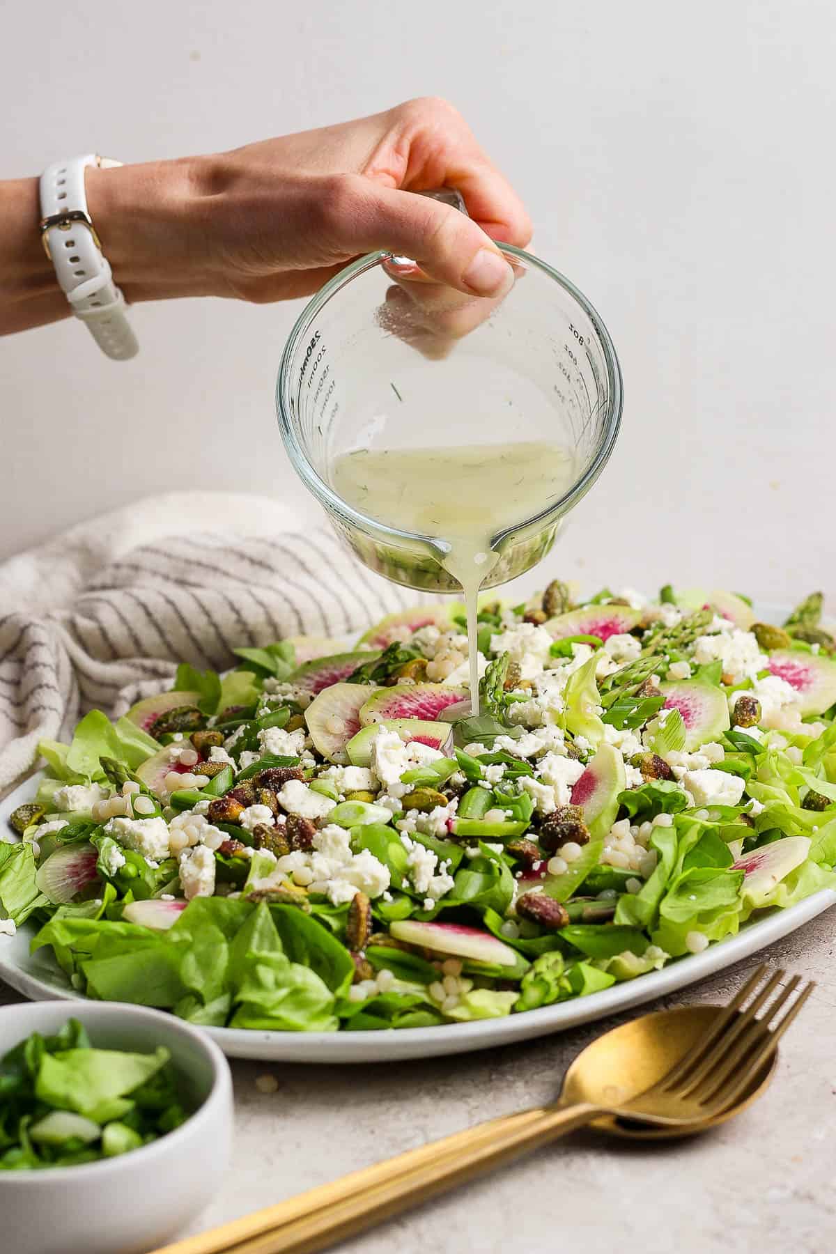 A homemade lemon dill vinaigrette being poured on a spring salad with couscous and peas next to a gold fork and spoon.