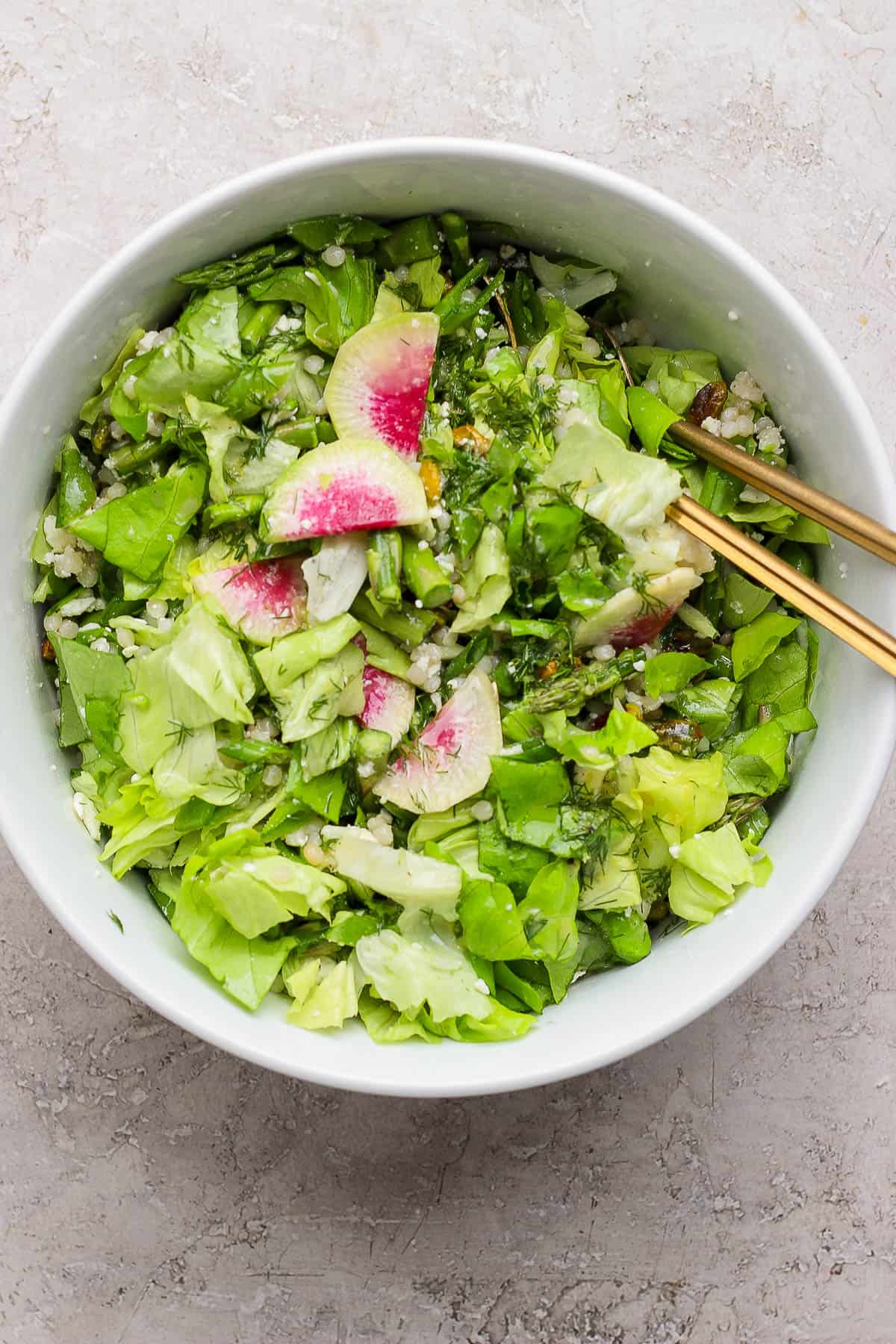 A spring salad with couscous and peas tossed together in a large bowl with gold serving utensils.