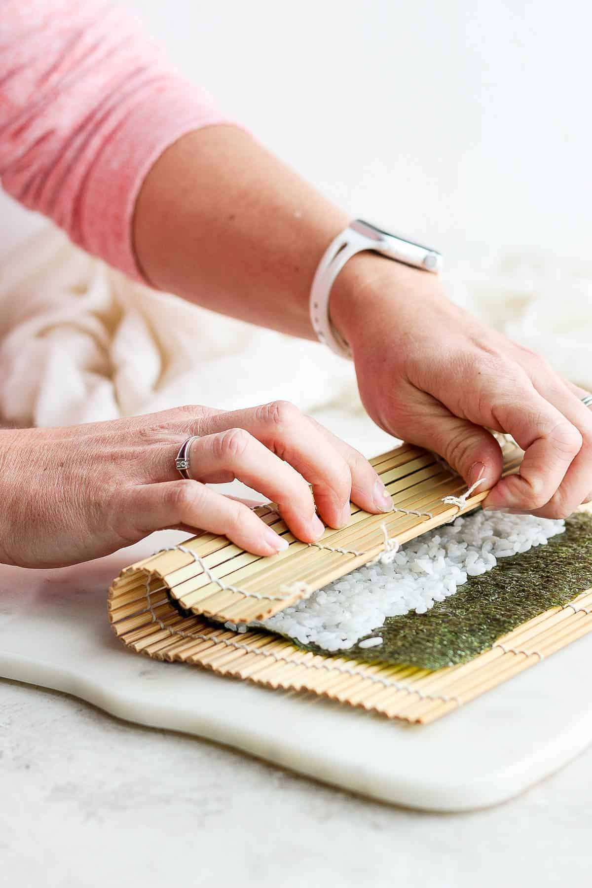 A bamboo mat being pulled over the beginning of a homemade sushi roll.