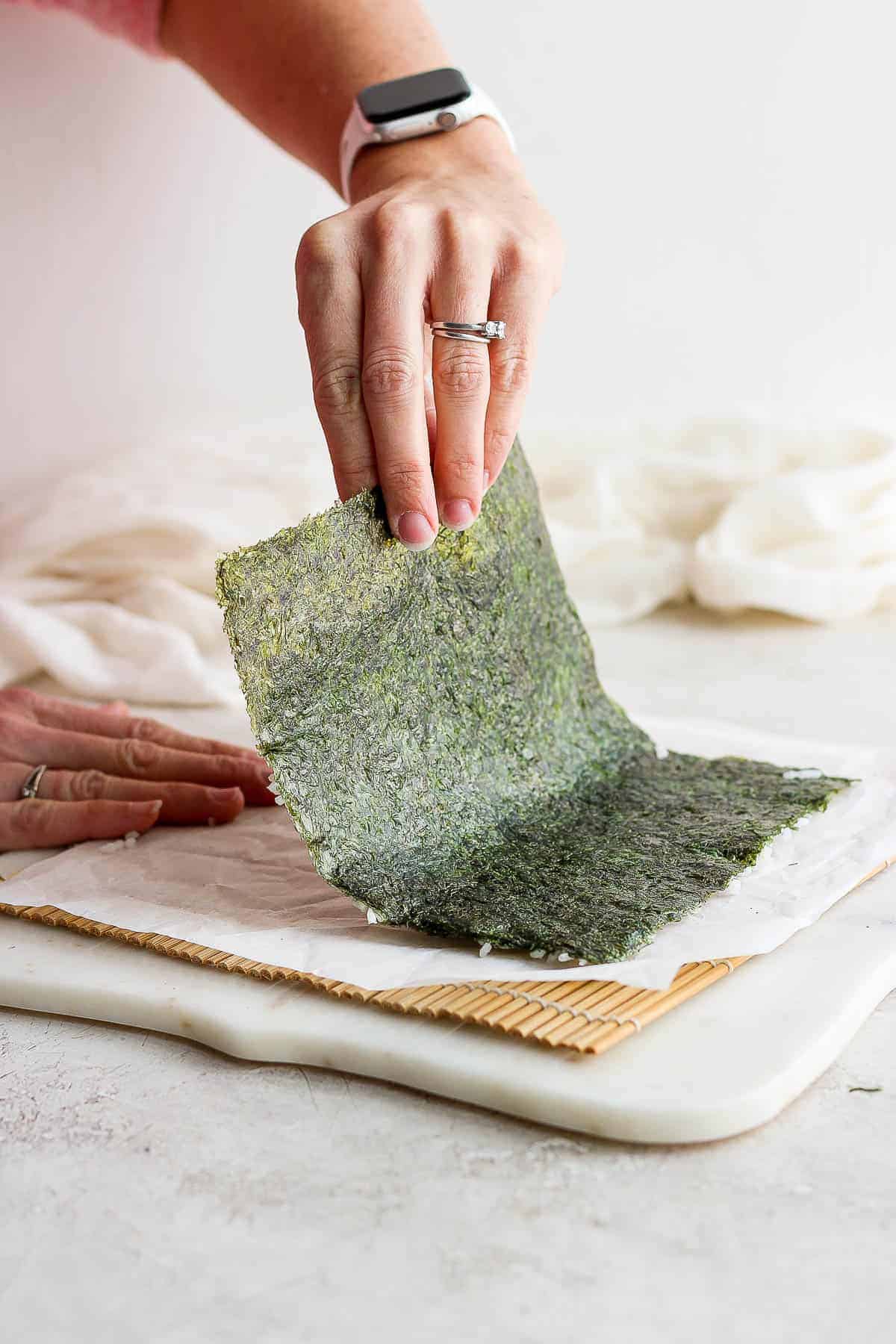 A rice-covered nori sheet being flipped over on top of a parchment-lined bamboo mat to make an inside out roll.
