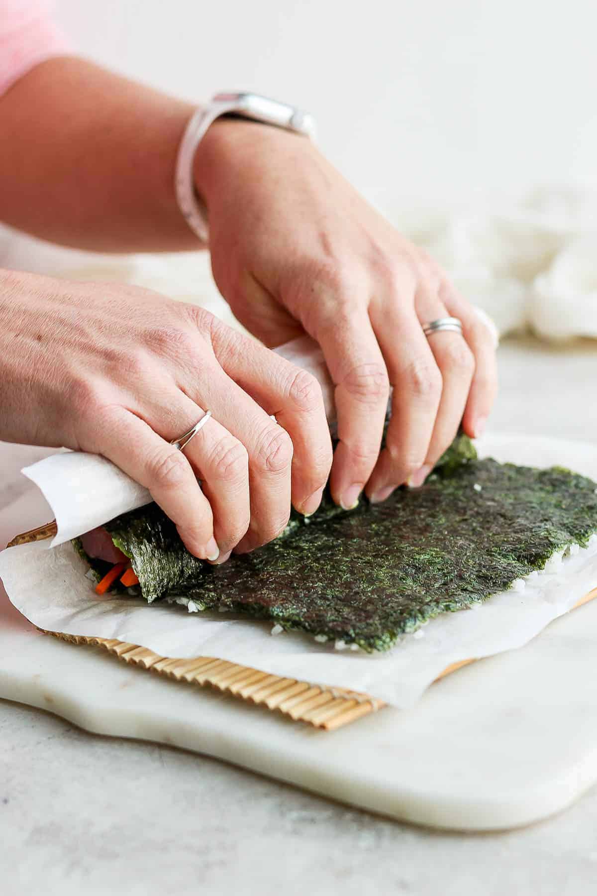 Two hands folding the end of a nori sheet, parchment paper and bamboo mat over the fillings of a sushi roll.