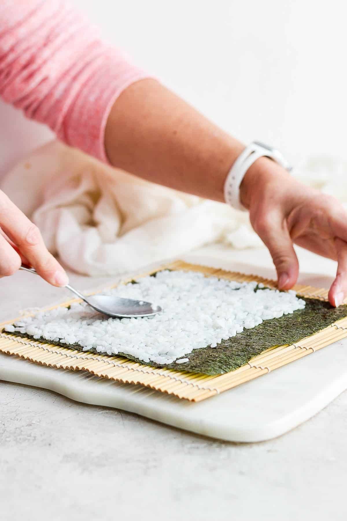Sushi rice being spread on a nori sheet with the back of a spoon for homemade sushi rolls.