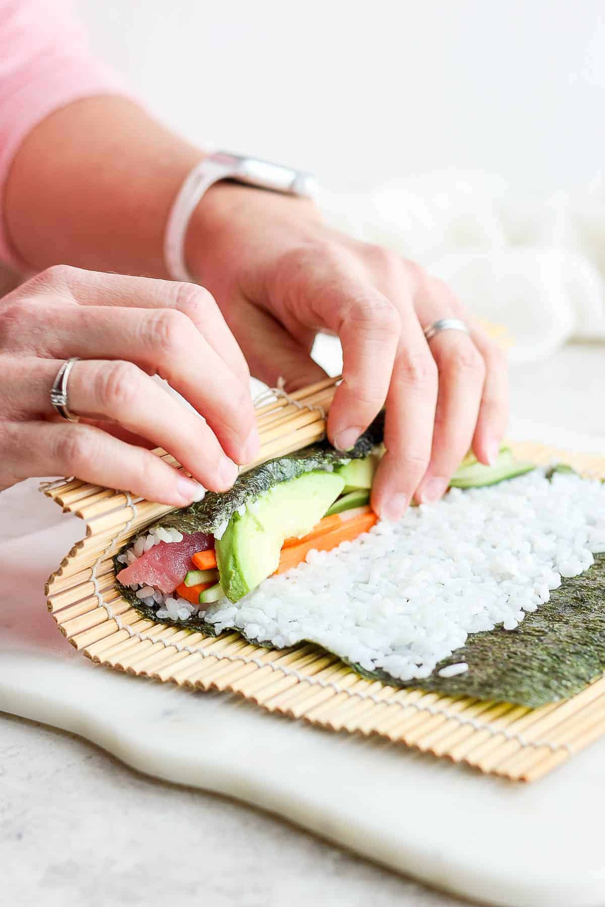 The edge of a nori sheet and bamboo sheet being lifted over the fillings of the sushi roll.