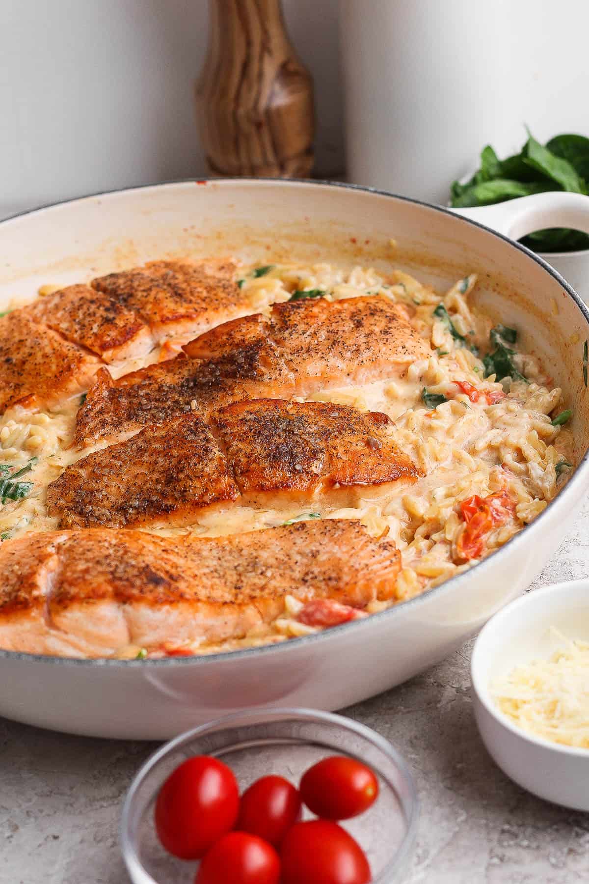 Salmon orzo in a white braiser next to small bowls of parmesan cheese and cherry tomatoes.