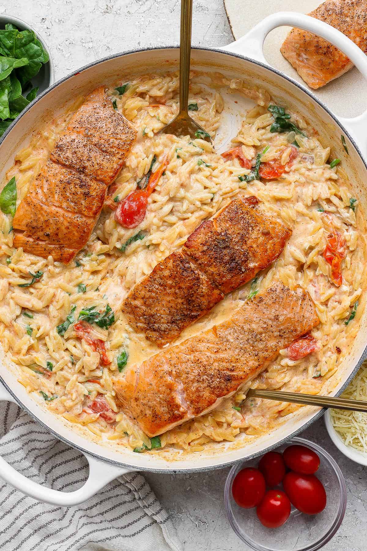 Salmon orzo in a large braiser with one salmon fillet removed and two gold spoons sticking out the sides.