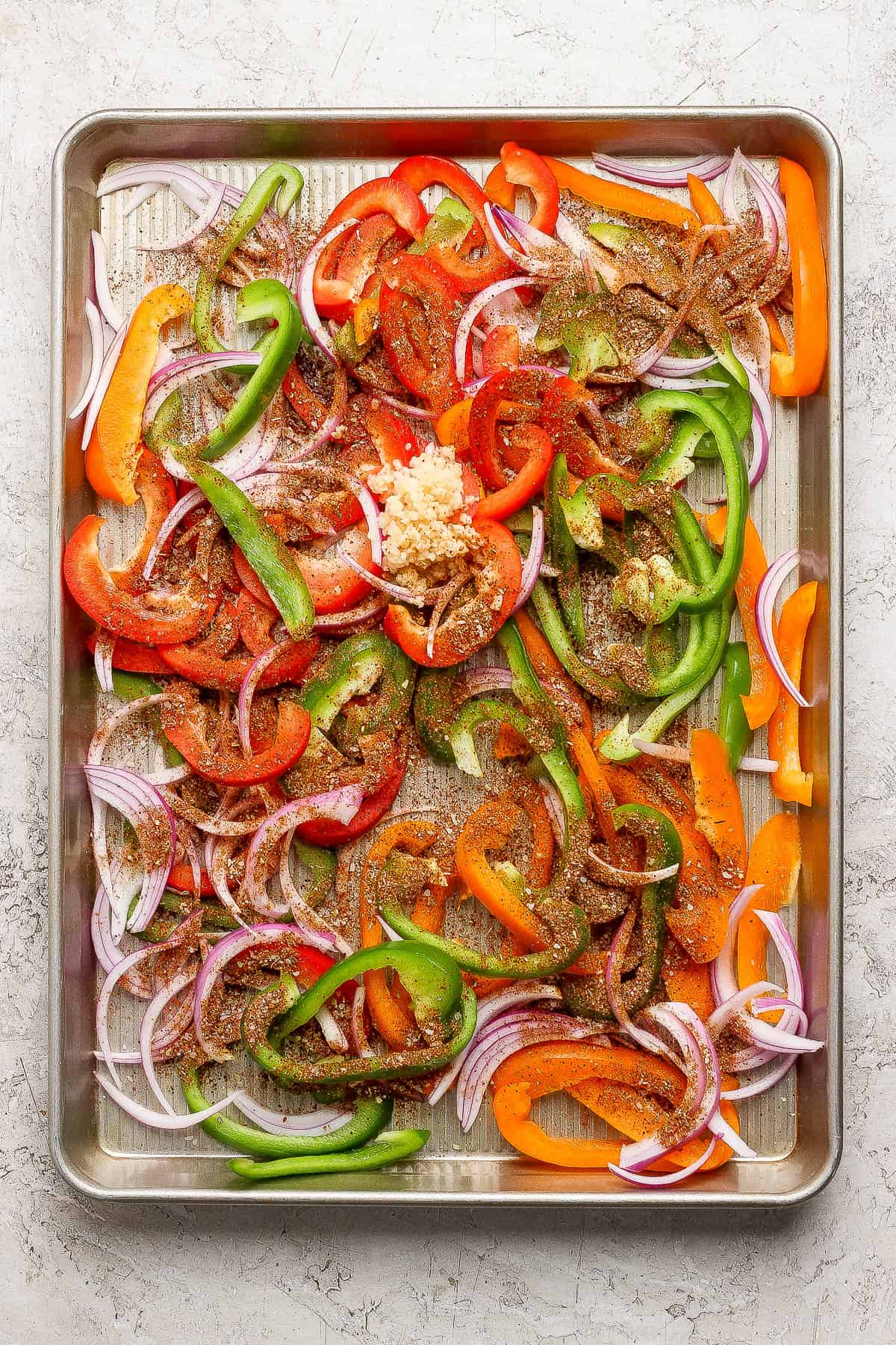 Sliced bell peppers and onions on a sheet pan with garlic and fajita seasoning.