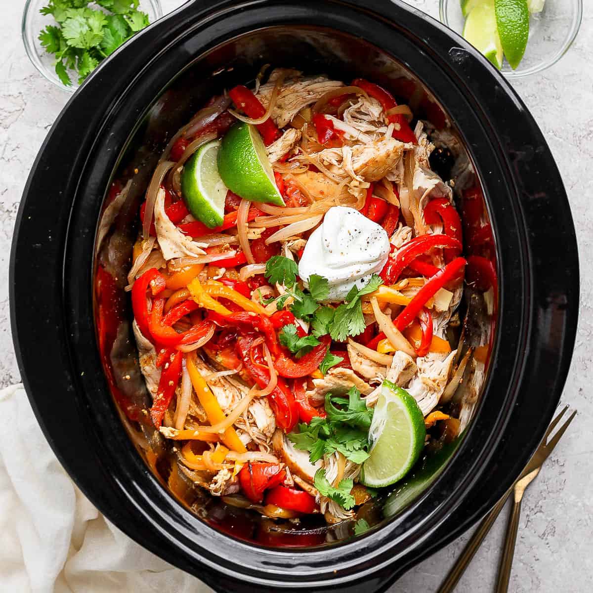Slow cooker chicken fajitas in the crockpot with some lime wedges and sour cream.
