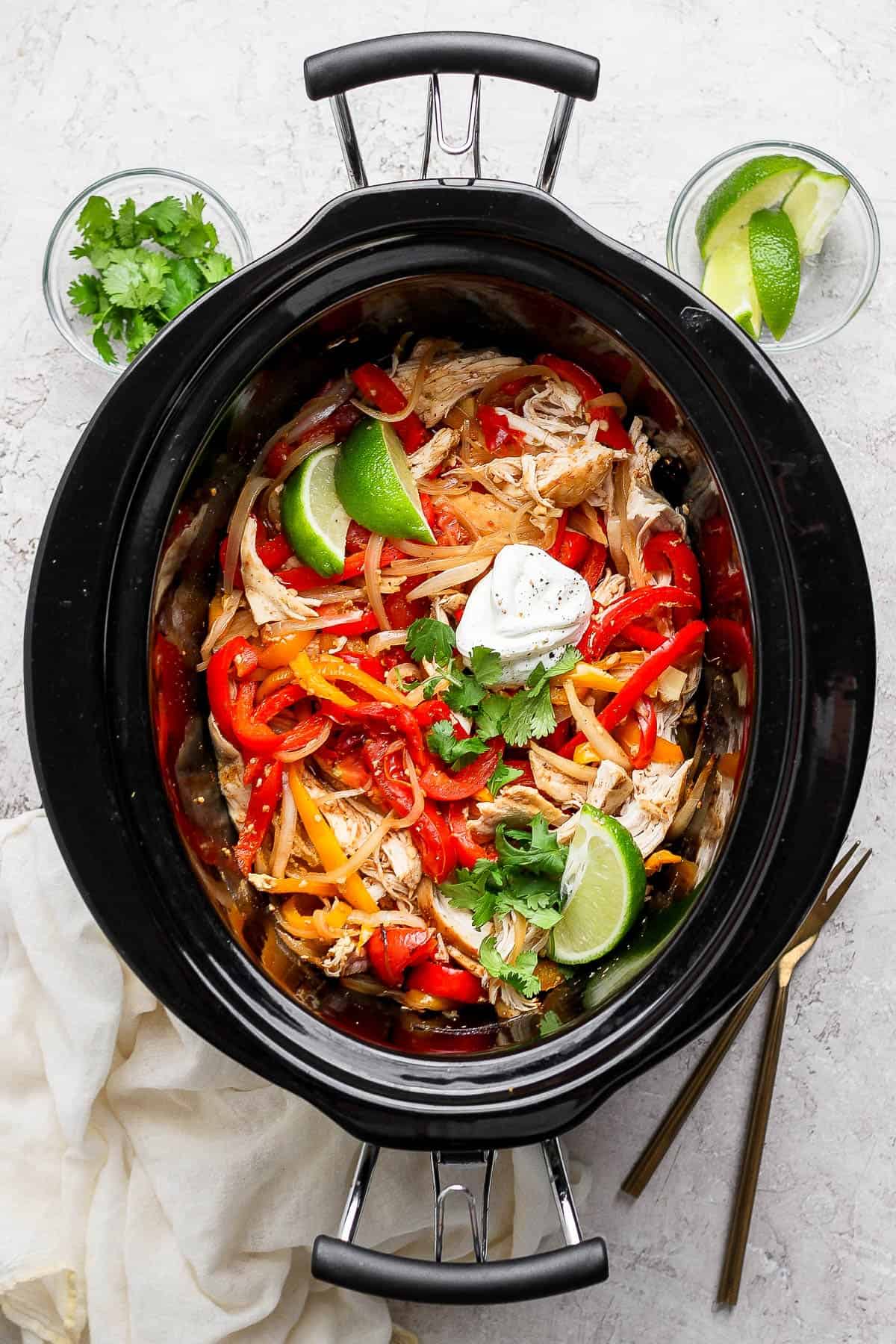 Slow cooker chicken fajitas in the crockpot with lime wedges, cilantro and sour cream.
