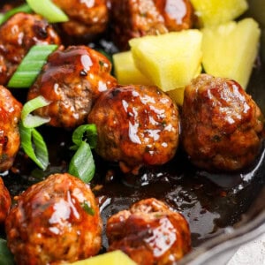 Pineapple teriyaki meatballs in a cast iron skillet with sliced green onions and pineapple chunks.