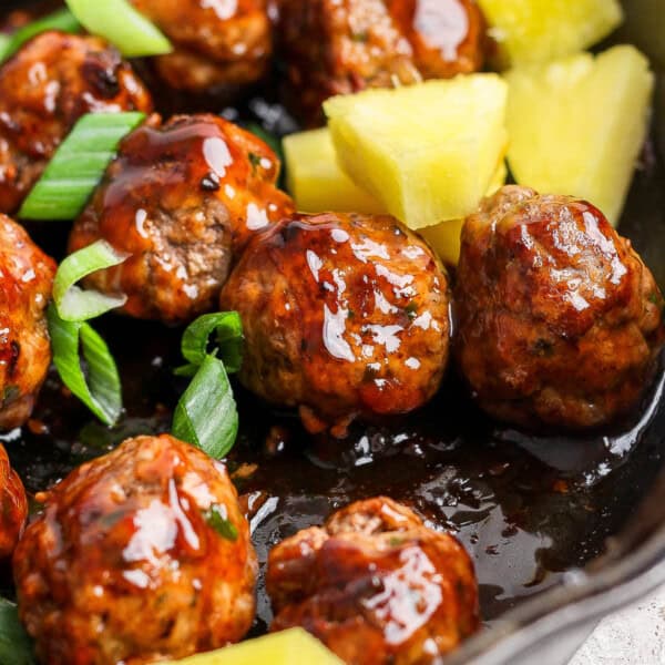 Pineapple teriyaki meatballs in a cast iron skillet with sliced green onions and pineapple chunks.