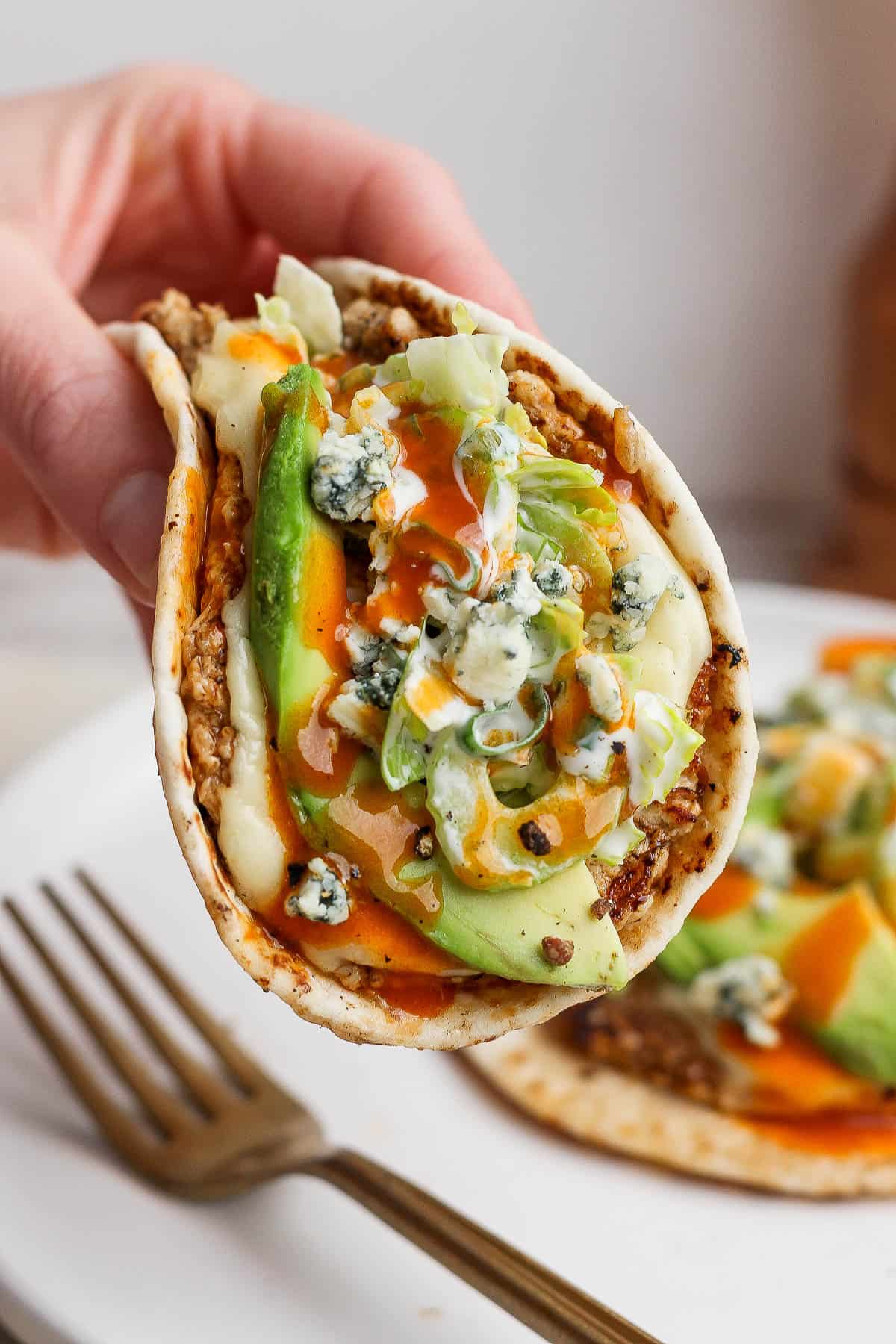 A hand holding a fully assembled buffalo chicken smash taco over another one on a plate with a fork.