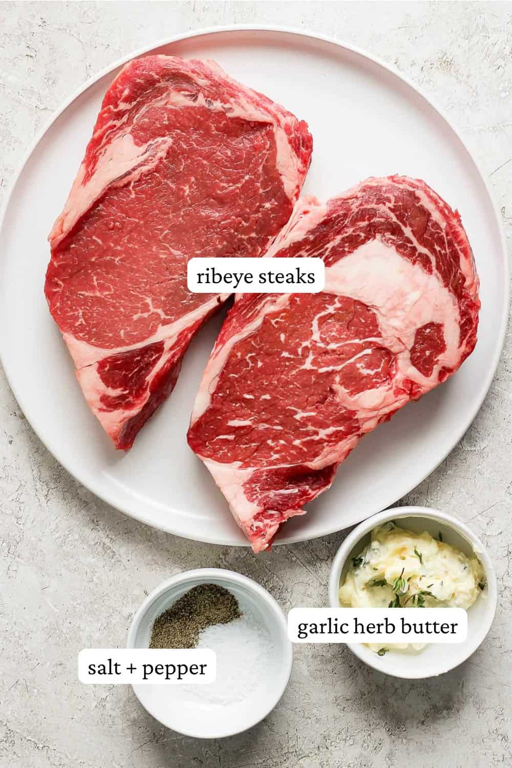 Ribeye steaks on a plate next to a bowl of salt and pepper and garlic herb butter with labels on them.