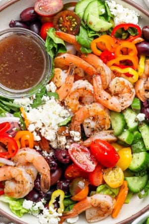 A fully assembled greek shrimp salad in a large bowl with a small dish of salad dressing.