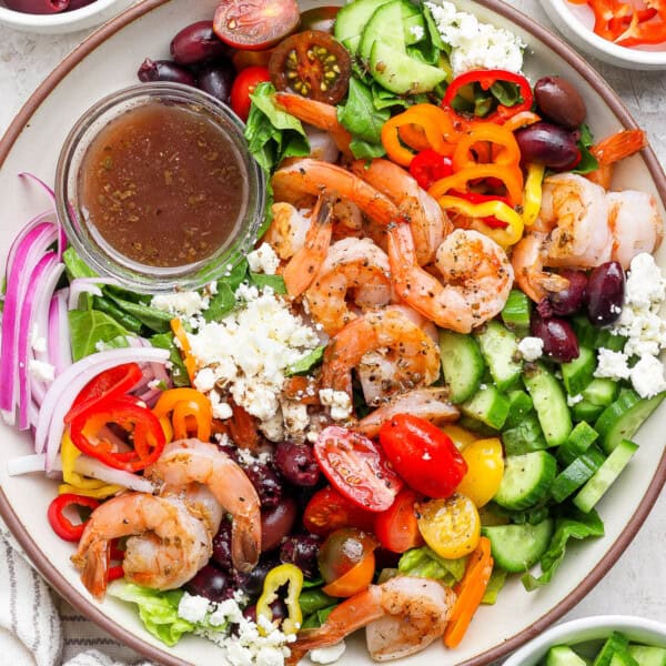 A fully assembled greek shrimp salad in a large bowl with a small dish of salad dressing.