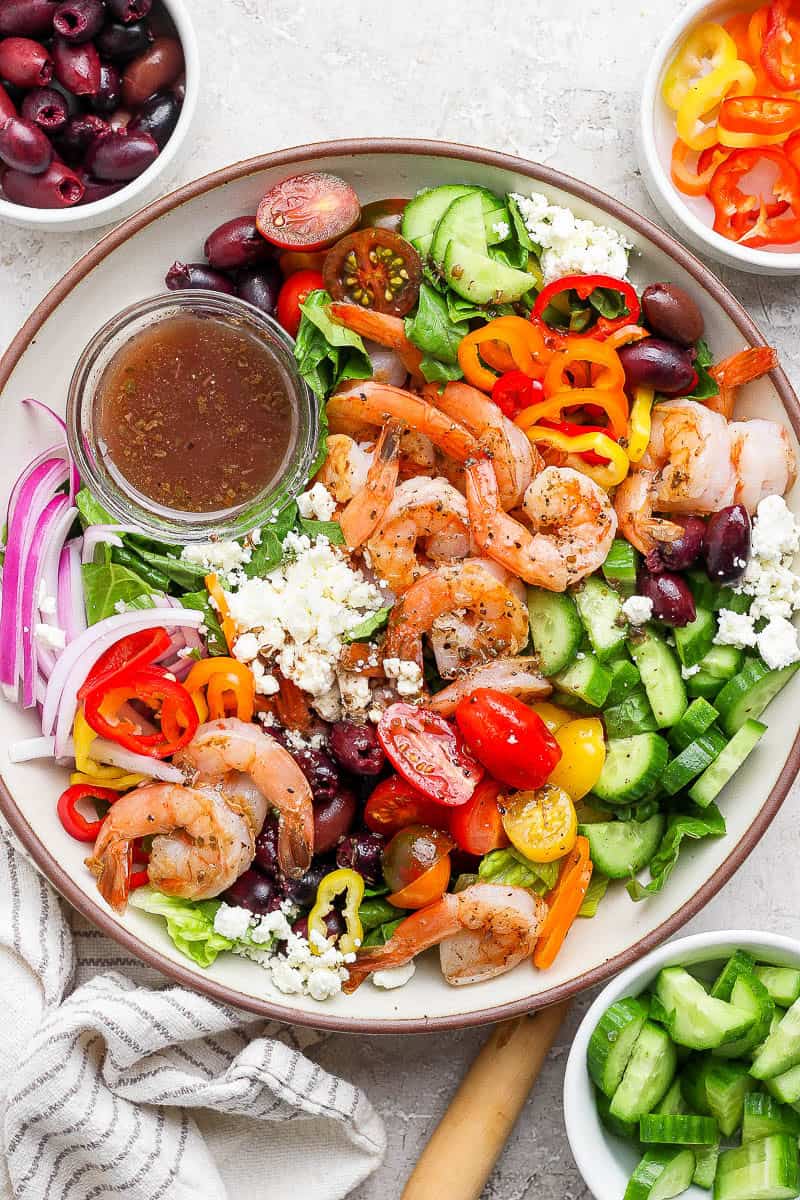 A fully assembled Greek shrimp salad next to small bowls of olives, sliced peppers, and cucumbers.