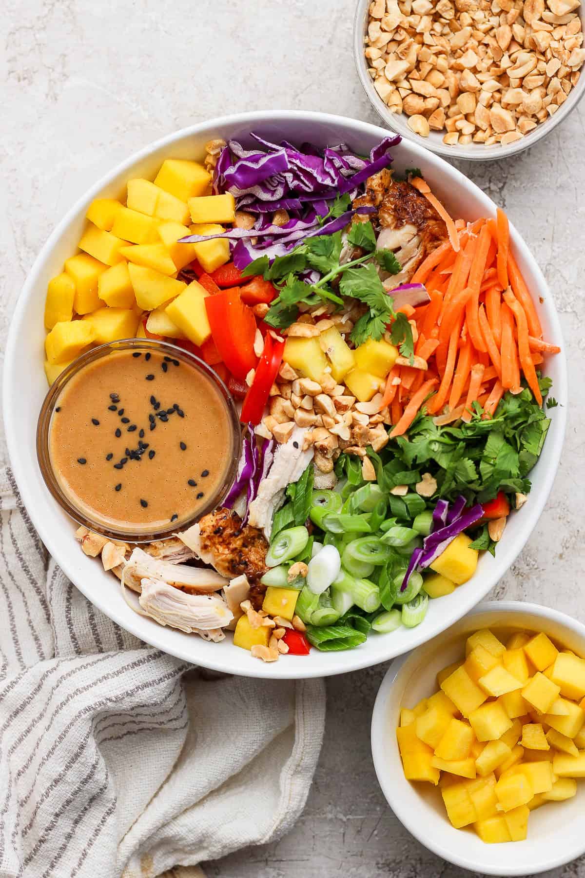 A fully assembled Thai peanut rotisserie chicken salad with a small dish of dressing sitting next to a bowl of peanuts and a bowl of mango.