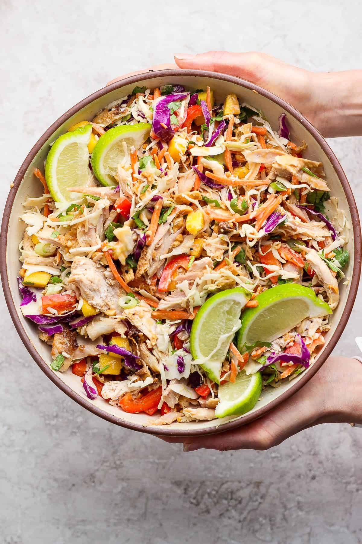 Two hands holding a large bowl of Thai peanut rotisserie chicken salad after it is all mixed together with the dressing.