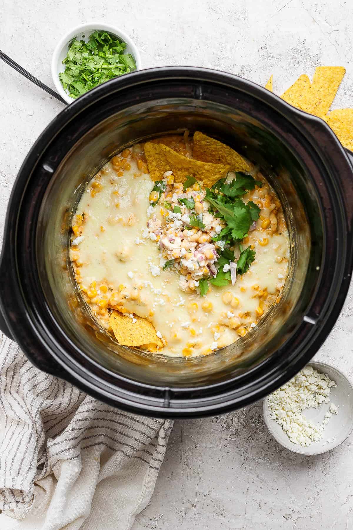 Street corn dip in a crockpot with cilantro and a couple tortilla chips next to small bowls of cilantro and feta cheese.