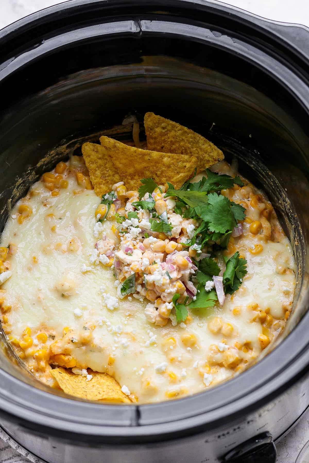 A couple tortilla chips and fresh cilantro on top of street corn dip in a slow cooker.
