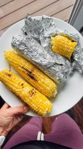 Two ears of Blackstone corn on the cob next to a fully wrapped ear of sweet corn and a cut piece of corn on the cob.